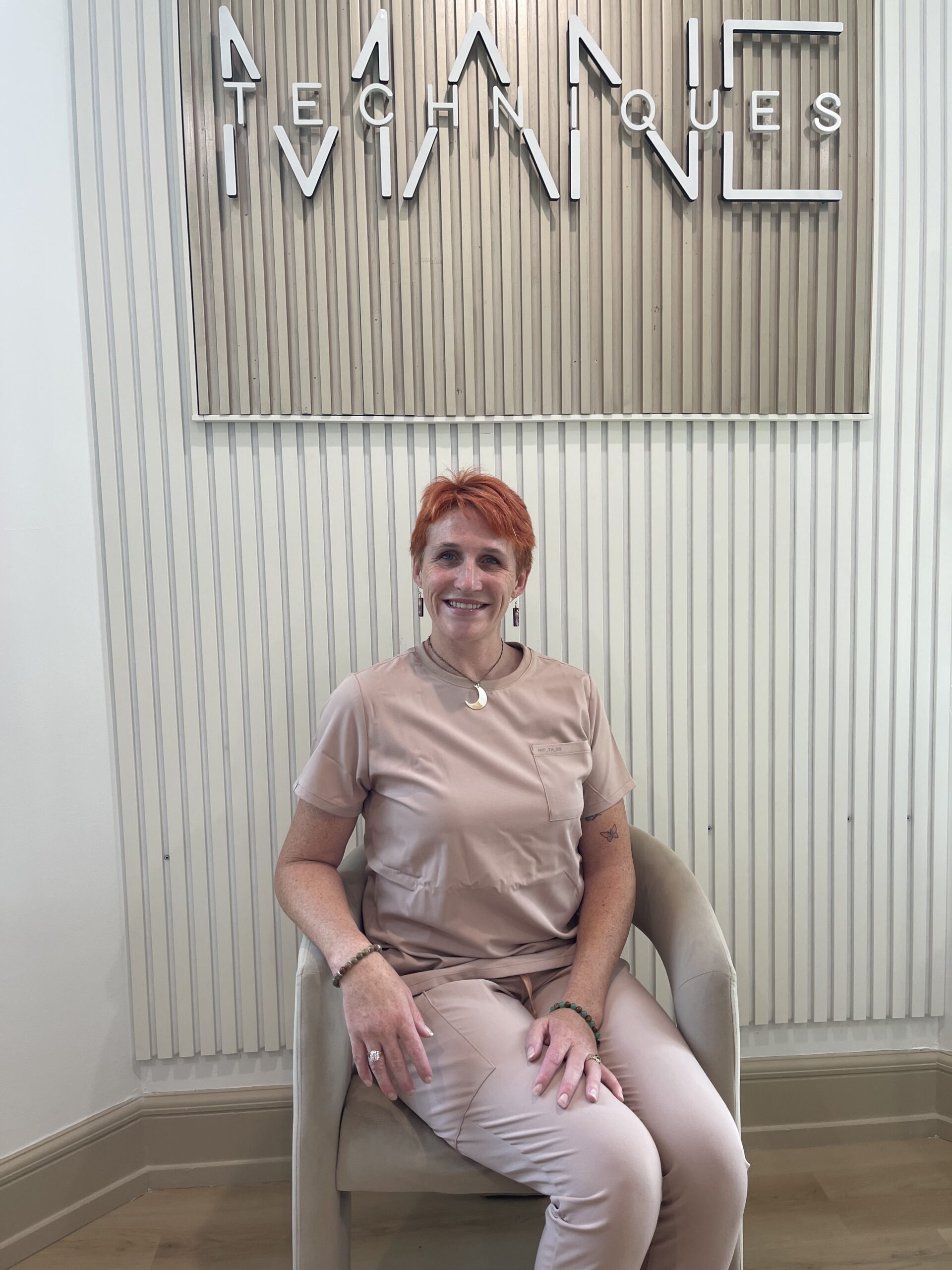 Smiling woman with short orange hair seated in a beige chair against a vertical striped wall with a large decorative sign behind her.