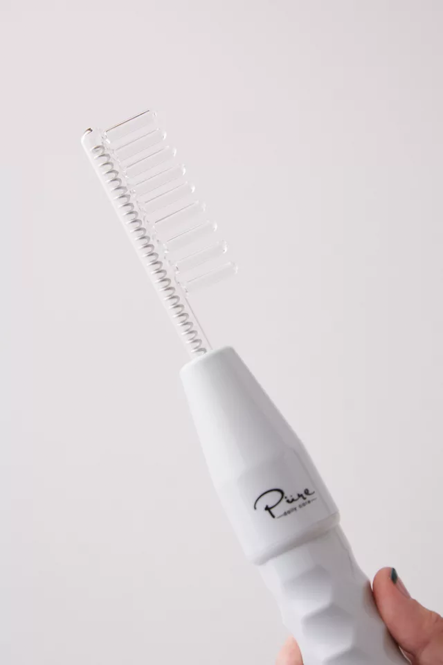 White heated hair styling brush with a row of comb-like teeth and a curved metal heating rod held by a hand, used for curling or smoothing hair.