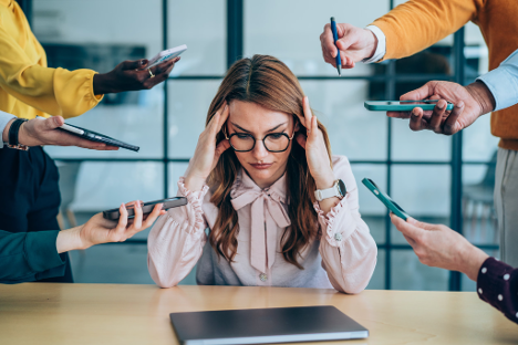 A stressed woman sits at a desk with hands on her temples as multiple people point phones and recorders toward her.