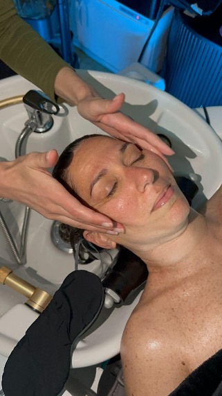 Woman lying back with eyes closed as a therapist massages her face/head at a facial treatment in a spa sink setup